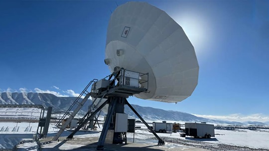A large satellite dish stationed in a snowy landscape with mountains in the background, under a clear blue sky.