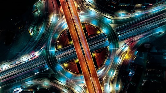 Aerial night view of a vibrant, multilayered traffic interchange with illuminated roads in swirling patterns of blue and orange lights.