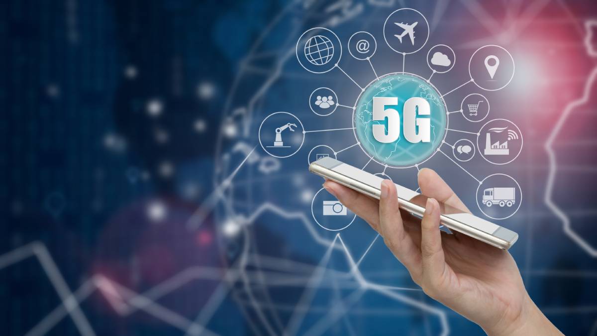 A hand holds a smartphone with a digital globe displaying '5G' surrounded by icons representing various connected technologies and applications, illustrating the concept of 5G network wireless systems and the Internet of Things.