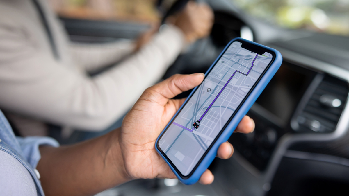 A close-up of someone holding a smartphone with a navigation app displayed on the screen, tracking the route of a vehicle while seated in a car.