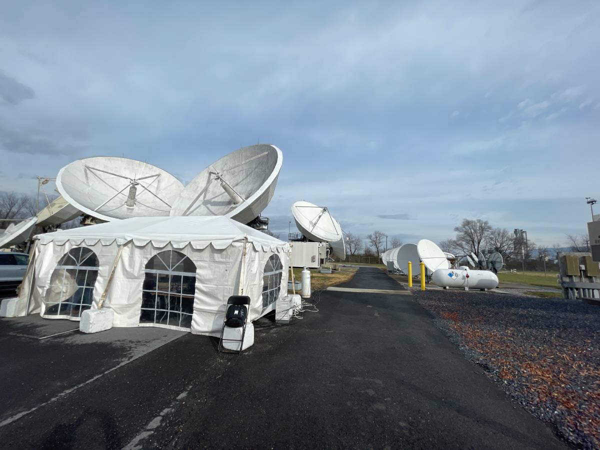 Large satellite dishes and a white tent are set up on a paved area for a virtual satellite communication demonstration.