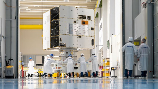 Technicians in cleanroom suits working on a large satellite module in a high-tech aerospace facility.
