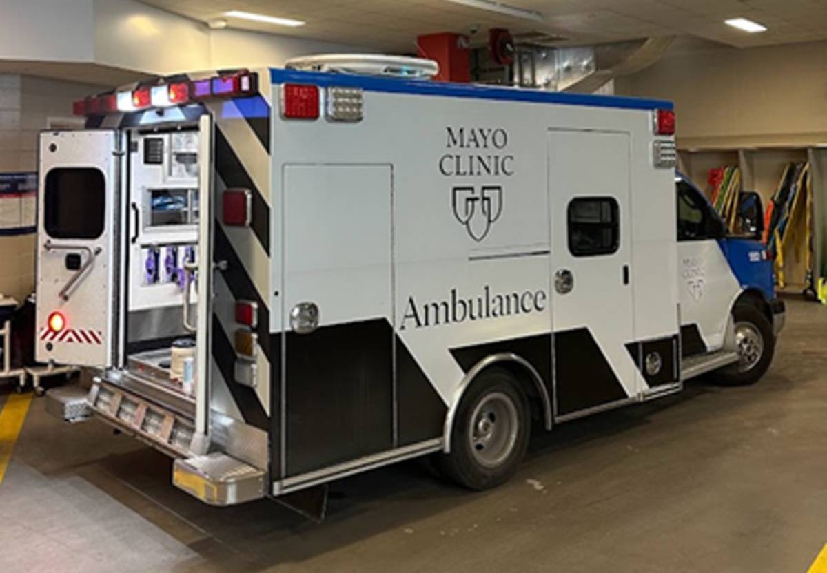 A Mayo Clinic ambulance parked inside a brightly lit garage facility with its rear door open, revealing interior medical equipment.