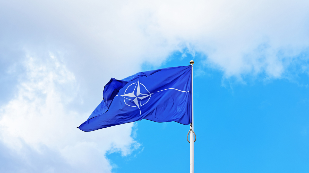 The NATO flag waving on a flagpole against a partly cloudy blue sky.