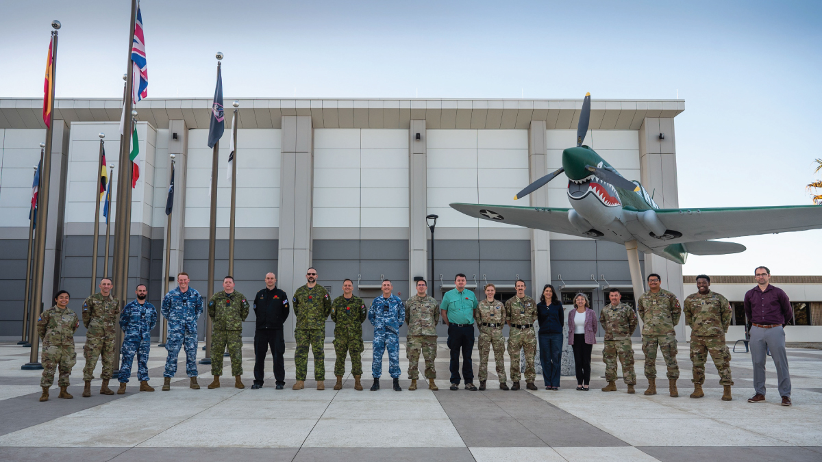 A group of military and civilian personnel stands in front of a building with international flags and a model of a fighter plane.