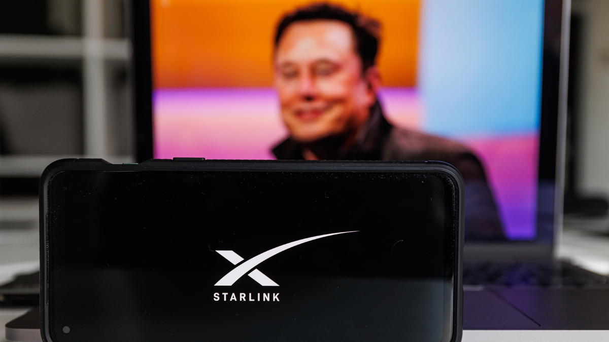 A Starlink router in the foreground with an out-of-focus image of Elon Musk on a laptop screen in the background.