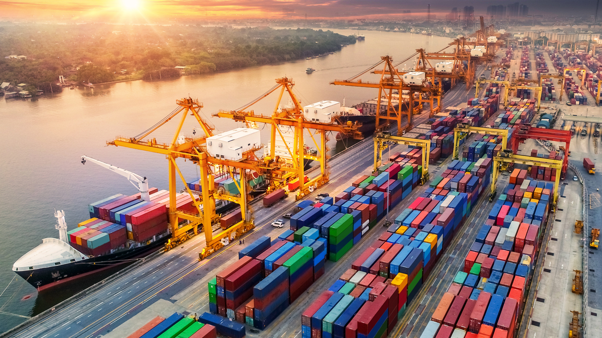 A bustling shipping port with numerous colorful cargo containers, cranes, and a docked cargo ship, set against a river and sunset backdrop.