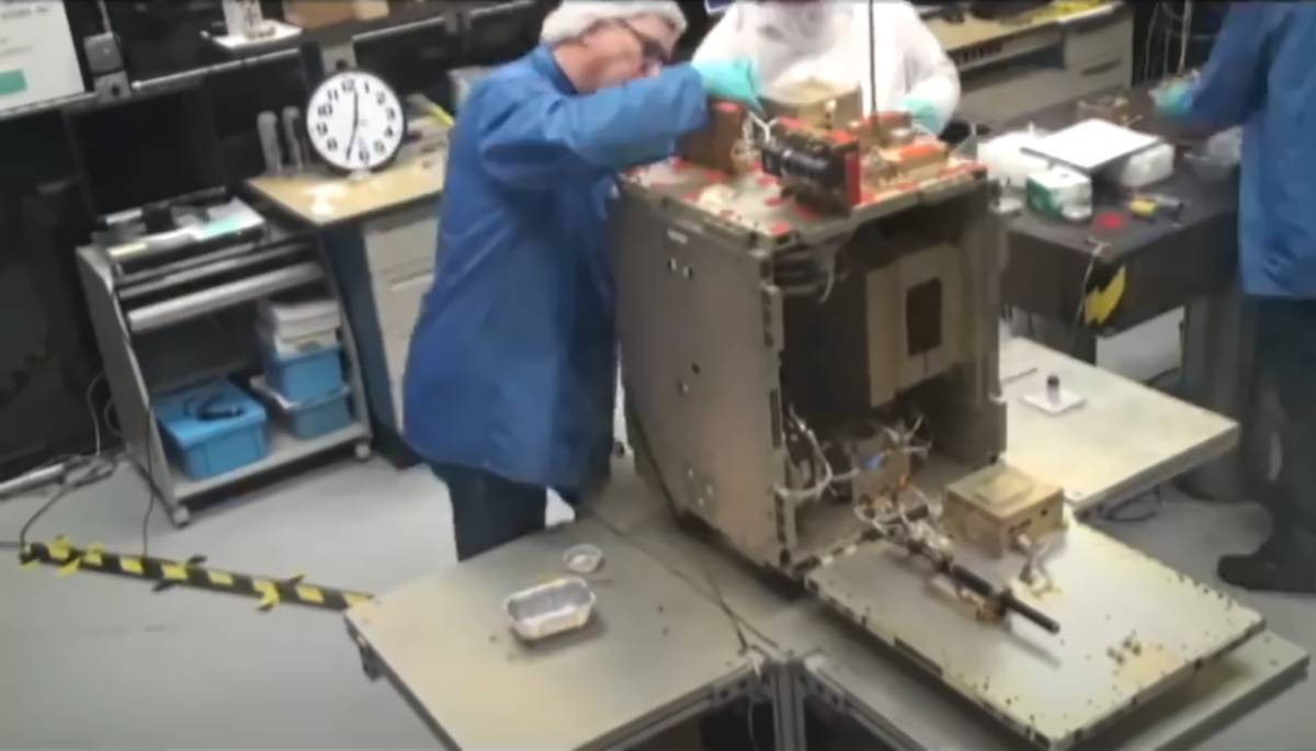 Technicians in a lab assembling or inspecting a satellite component on a workbench, surrounded by equipment and tools.