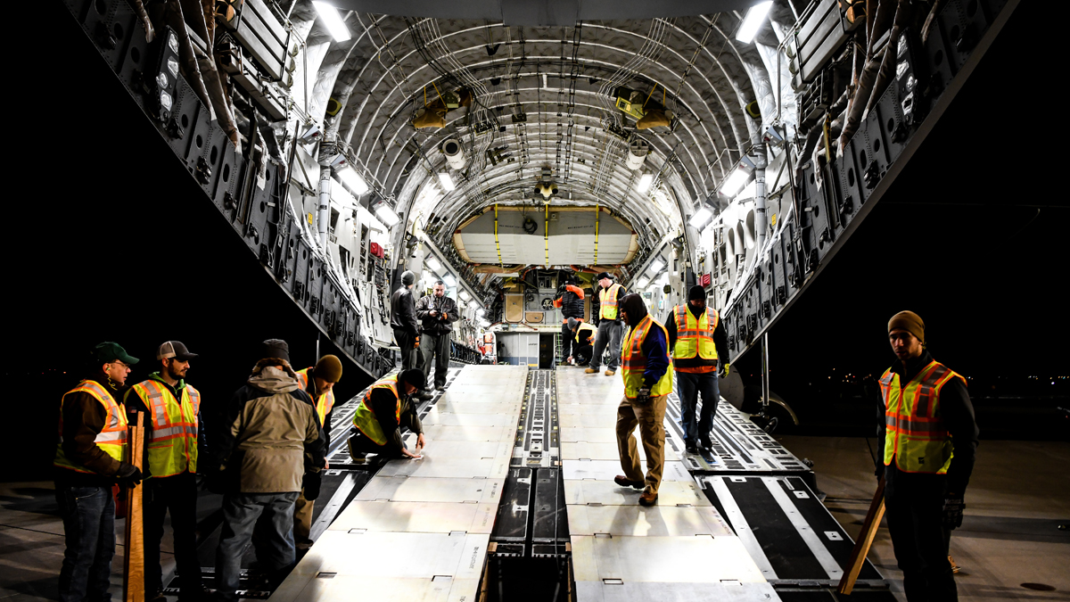 Workers inside a large cargo aircraft prepare and inspect equipment during a nighttime operation, wearing high-visibility vests and surrounded by industrial lighting.