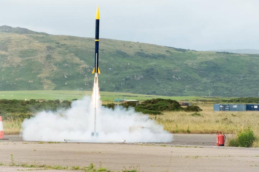 A rocket with a blue and yellow color scheme, designed and built by the Durham University team, is shown launching from a pad with smoke billowing around it, set against a backdrop of grassy hills and a cloudy sky.