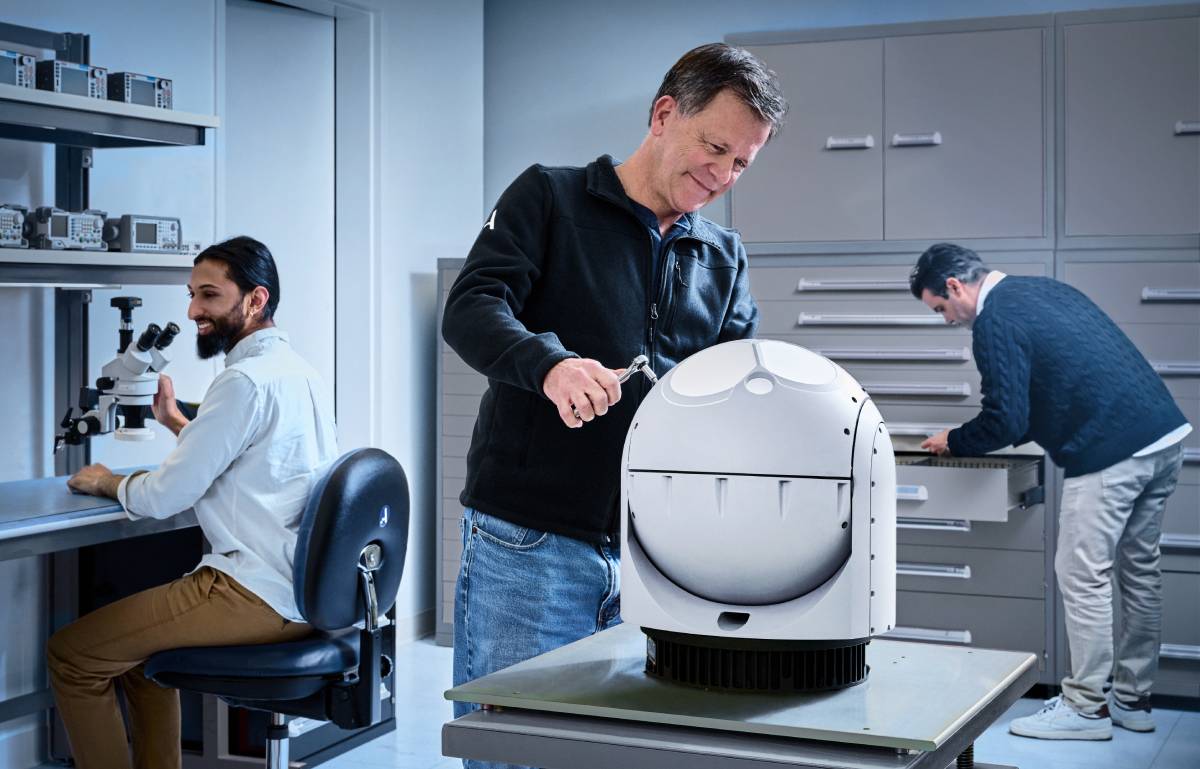 Three engineers are working in a laboratory setting, with one man assembling a large, spherical device, another inspecting components in a drawer, and a third using a microscope, all surrounded by technical equipment.