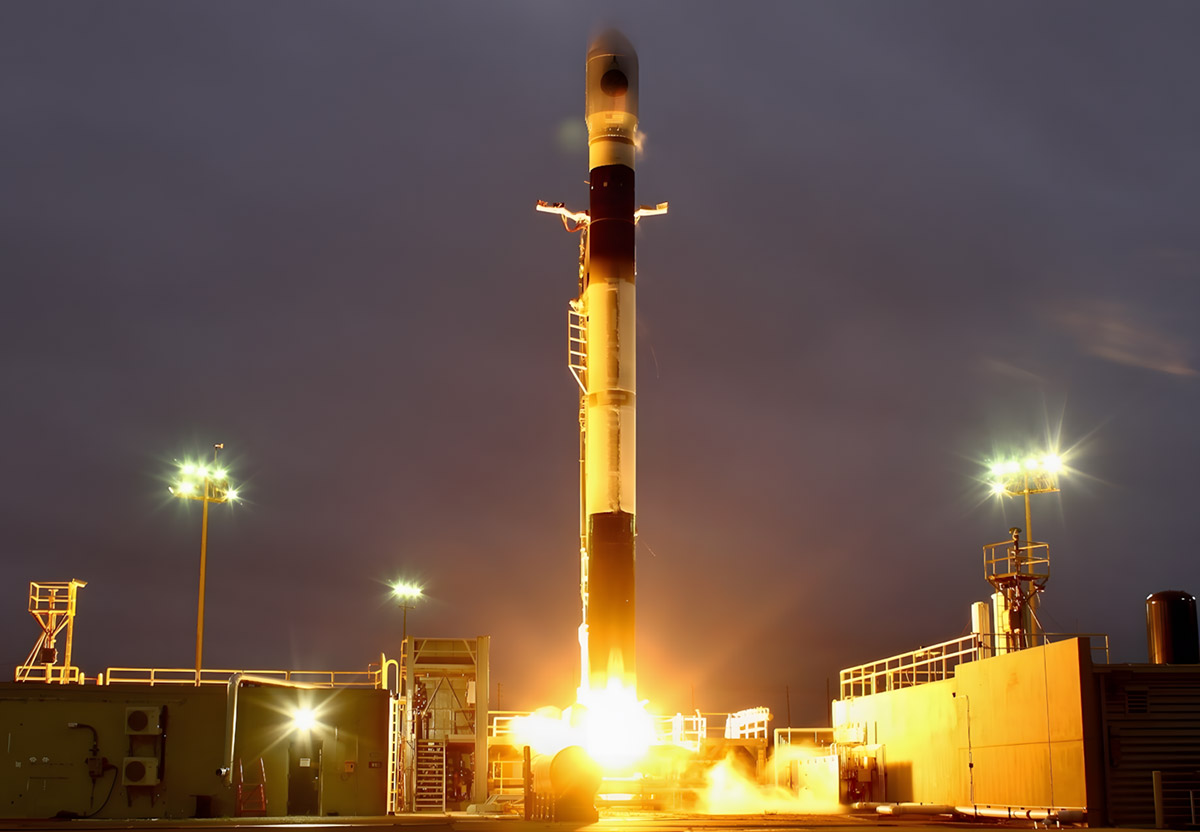 A rocket launches into the night sky, emitting bright flames, with a structured platform and lighting around the launch site.