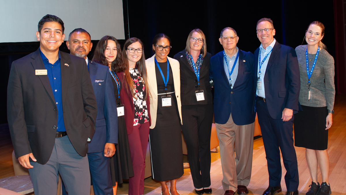 A group of professionals standing together, smiling for a photo, with conference badges and lanyards around their necks.