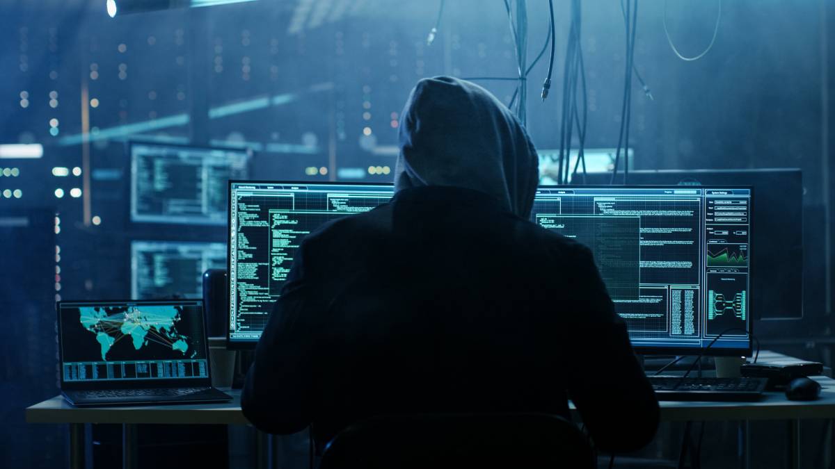 A person wearing a hoodie is sitting in front of multiple computer screens displaying code and data in a dimly lit room.