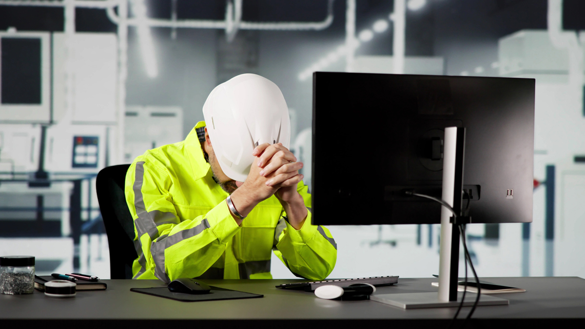 A worker in a high-visibility jacket and hard hat sits at a desk in front of a computer, leaning forward with their head in their hands, suggesting stress or fatigue.