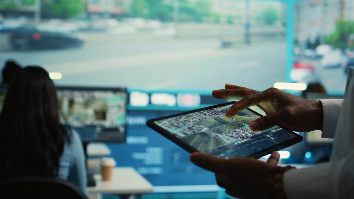A person is using a tablet with a detailed map interface, while others work on computers in a control room with a large screen showing a blurry outdoor scene in the background.