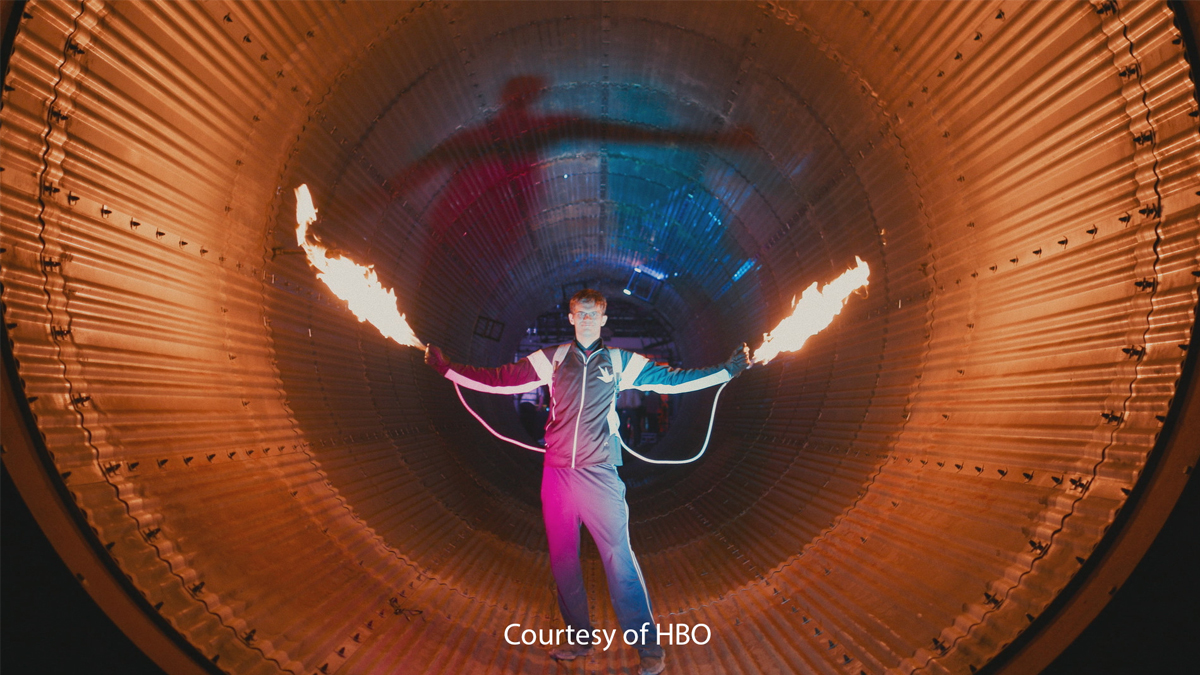 A person stands inside a large cylindrical tunnel, holding two flaming torches with arms outstretched, surrounded by vibrant orange and blue lighting.