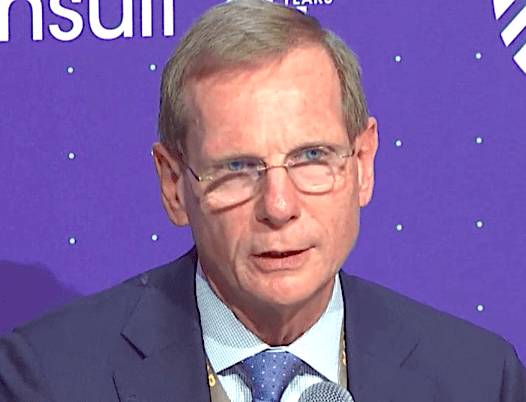 A man wearing glasses and a suit speaks at a conference, with a purple backdrop behind him.