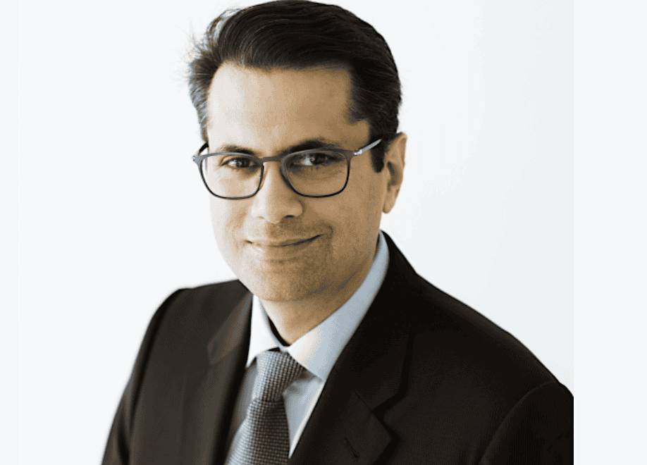 A man wearing glasses and a suit smiles slightly while posing for a professional headshot against a light background.