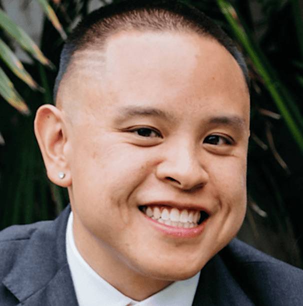 A smiling individual wearing a suit, posing outdoors with greenery in the background.