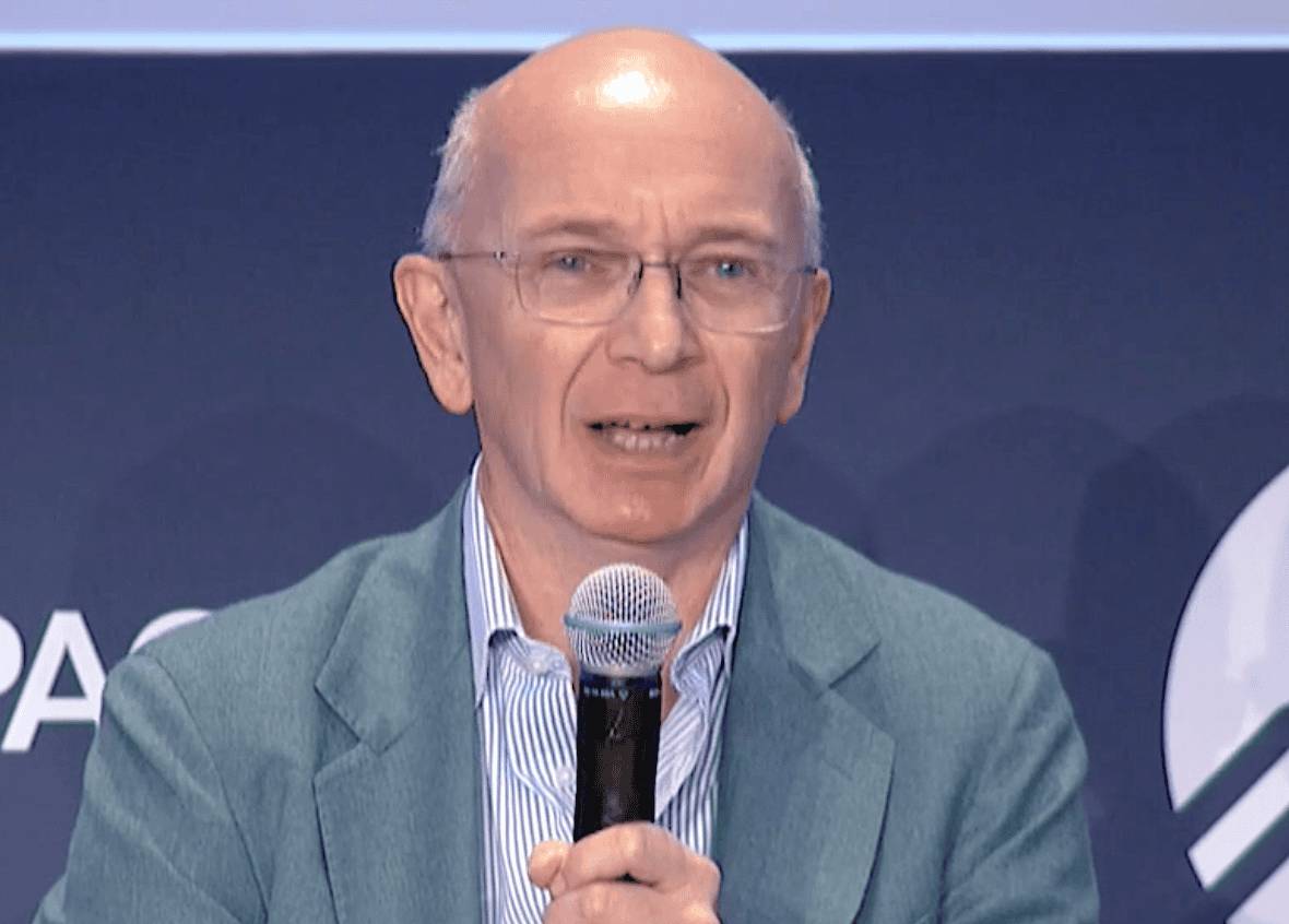 A man in glasses and a green suit speaks into a microphone at a formal event.