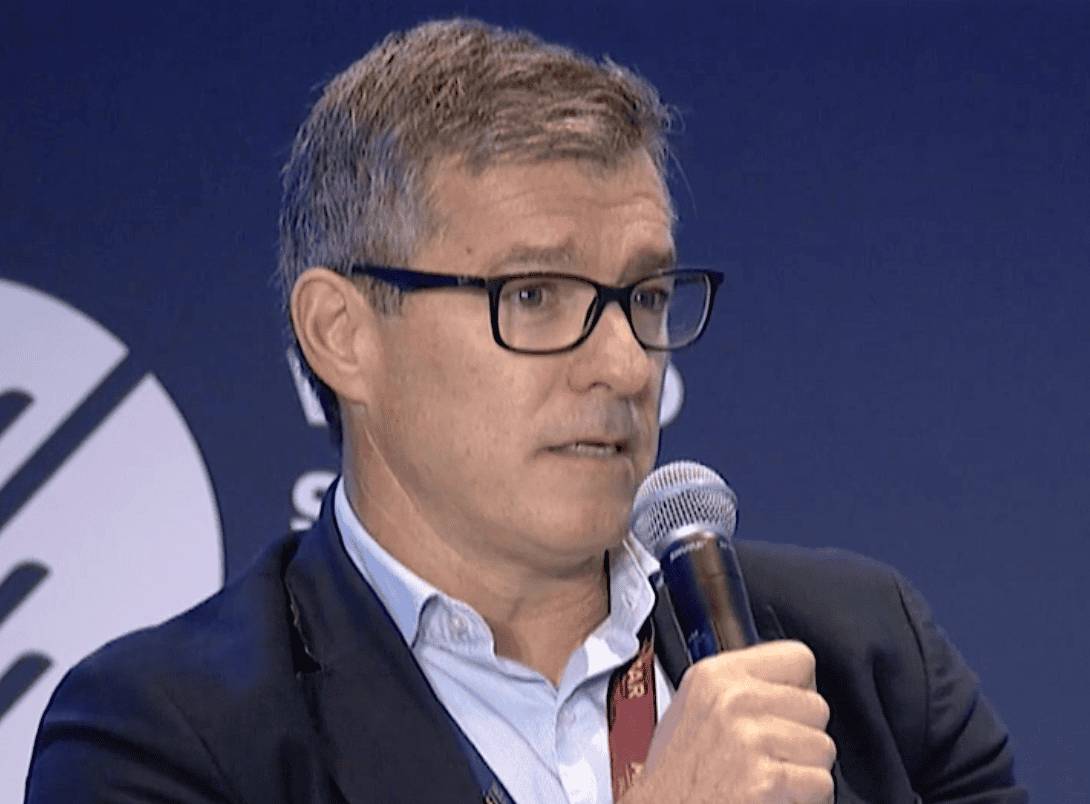 A man with glasses and a jacket is speaking into a microphone during what appears to be a formal panel or discussion.