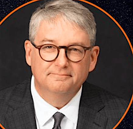 A man with short gray hair and glasses, dressed in a suit and tie, posing for a professional headshot against a dark background.