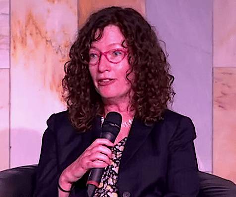 A woman with curly hair and glasses, wearing a dark jacket, holding a microphone while speaking at an event.