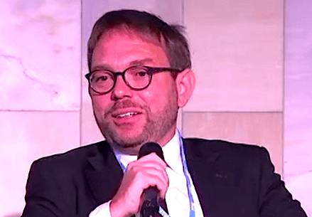 A man with short hair and glasses, wearing a suit and tie, holding a microphone while speaking at an event.