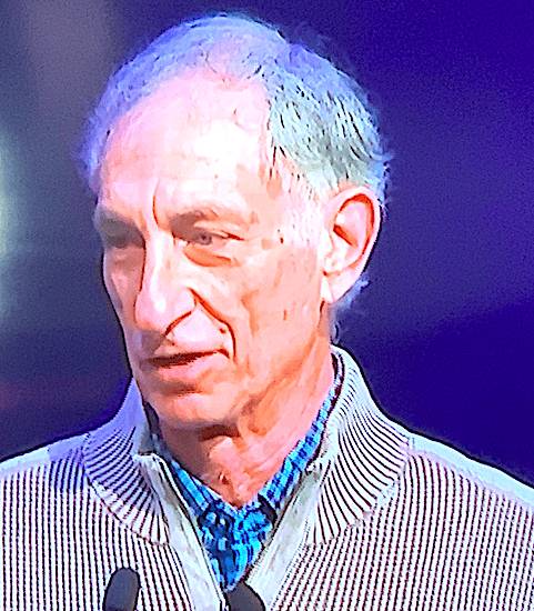 A close-up of an older man with gray hair wearing a light-colored jacket over a blue collared shirt, speaking on stage with a microphone in front of him.