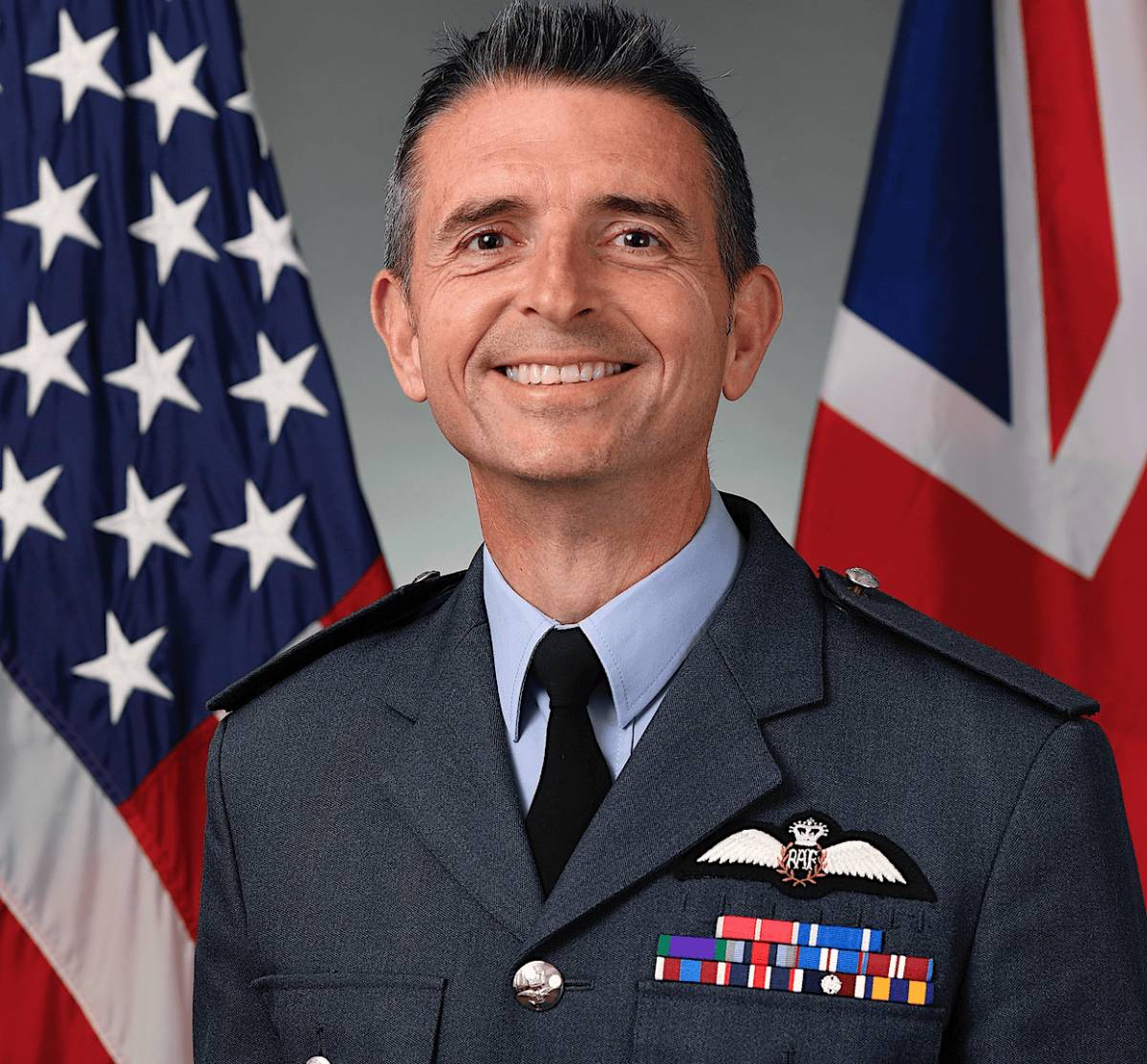 Smiling military officer in formal uniform with RAF wings insignia, standing in front of American and British flags.