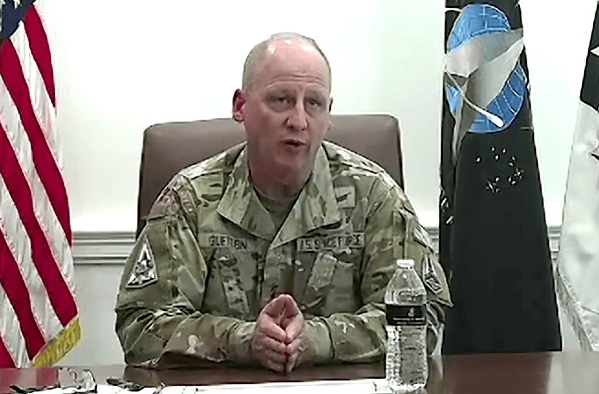 Military officer in camouflage uniform seated at a table, with American and Space Force flags in the background, addressing an audience.