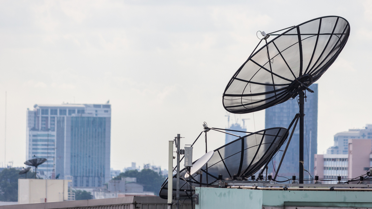 Damaged satellite dishes and antennas on the rooftop of an old building, with a city skyline in the background.