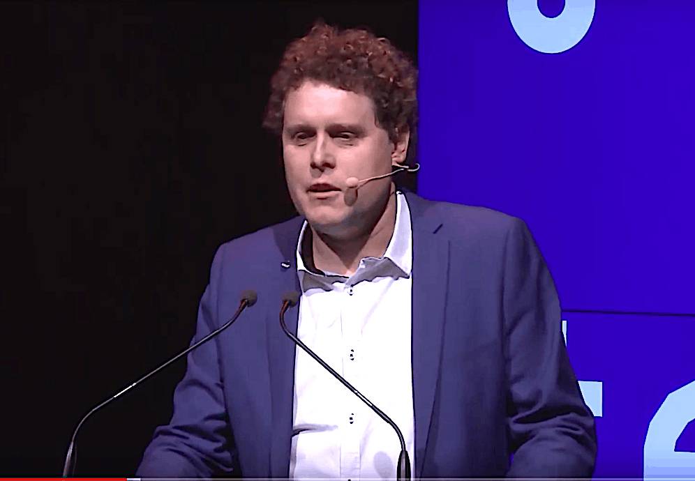 A man with curly red hair in a blue suit speaks into a headset microphone on stage, standing behind two microphones with a purple screen in the background.