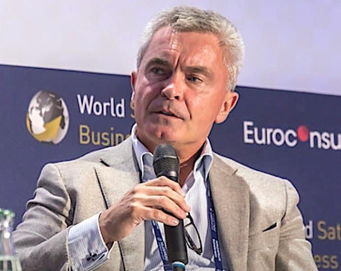 A man in a light gray blazer speaking into a microphone at a business conference, with banners in the background reading 'World Satellite Business Week' and 'Euroconsult.'