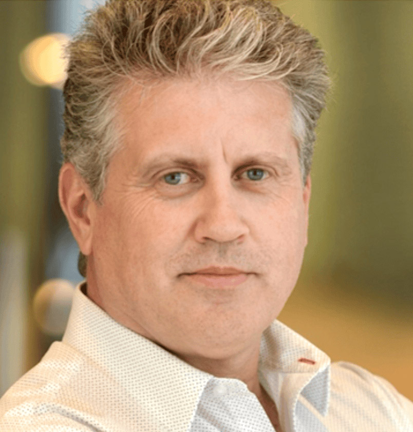 A portrait of a man with short, wavy gray hair wearing a white collared shirt, set against a softly blurred background.