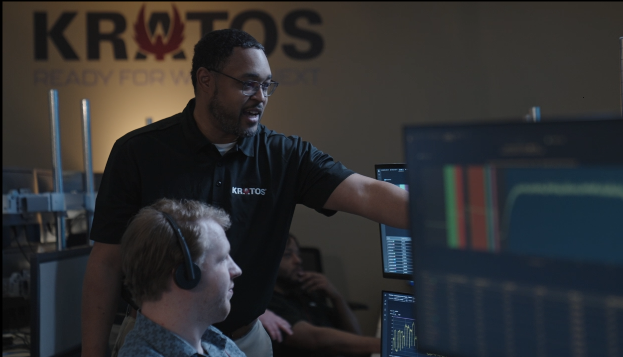 A man in a Kratos shirt points to a computer monitor while assisting a colleague wearing a headset in a control room, with multiple screens displaying data charts and graphs.