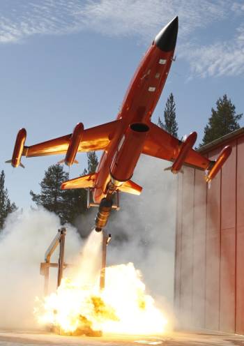 A bright orange unmanned aerial vehicle (UAV) launching vertically from a platform, with flames and smoke erupting beneath it, likely indicating the use of a rocket booster for takeoff. The UAV is a BQM-167i, which is used for aerial target practice or training exercises.