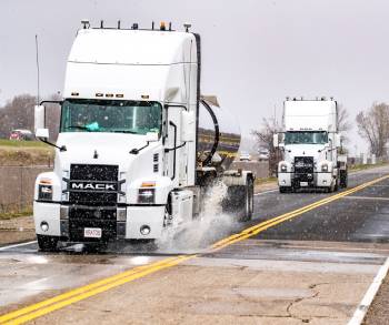 Two white Mack trucks are driving in tandem on a wet road, splashing water, with the front truck leading the second truck in a Leader Follower Platooning formation.