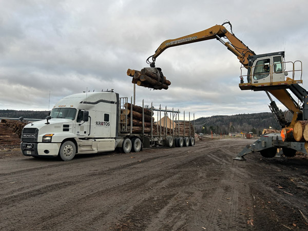 A white Mack semi-truck is loaded with timber logs using a large yellow hydraulic loader at a lumber yard under an overcast sky.